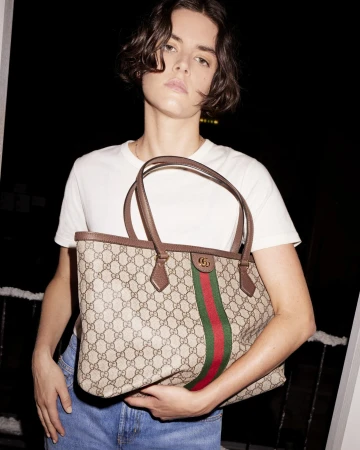 Adult female wearing a white shirt and blue jeans, holding a Gucci tote bag with red and green stripes in an indoor setting.