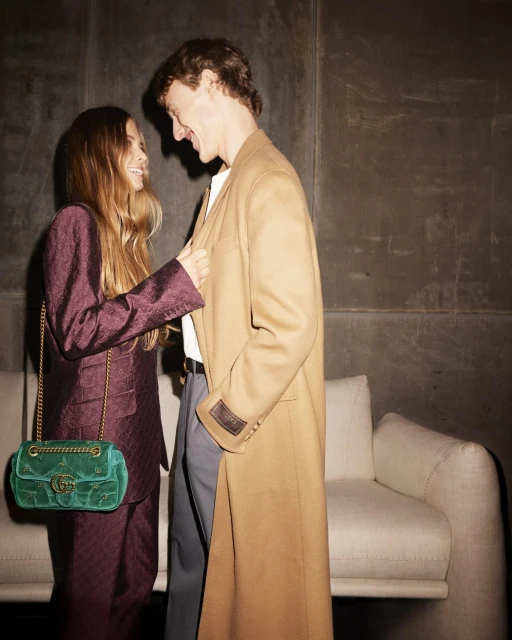 A stylish couple in an indoor setting. The woman wears a purple suit with a green velvet purse and the man wears a tan overcoat.