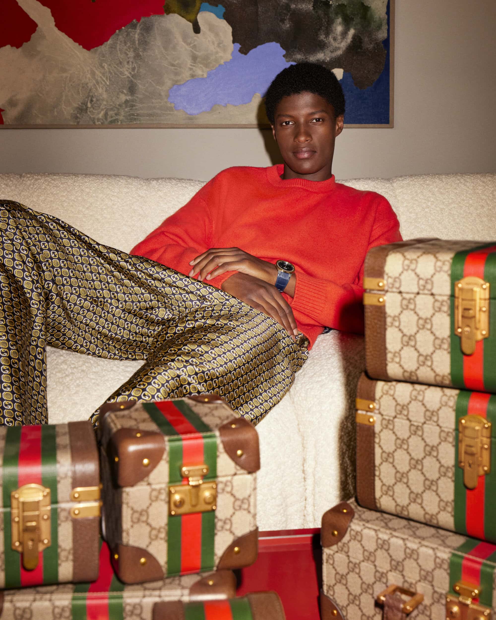 Young man in red sweater and patterned trousers seated on a white sofa surrounded by monogram vanity cases for Gucci Holiday 2023