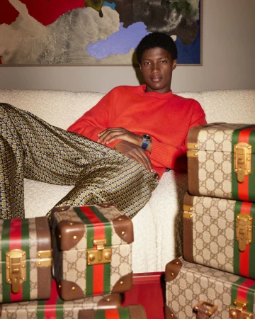 Young man in red sweater and patterned trousers seated on a white sofa surrounded by monogram vanity cases for Gucci Holiday 2023