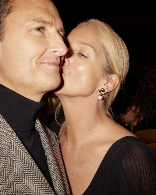 Close-up of a Caucasian woman kissing a Caucasian man on the cheek, both with light hair, at an indoor event with a dark background.