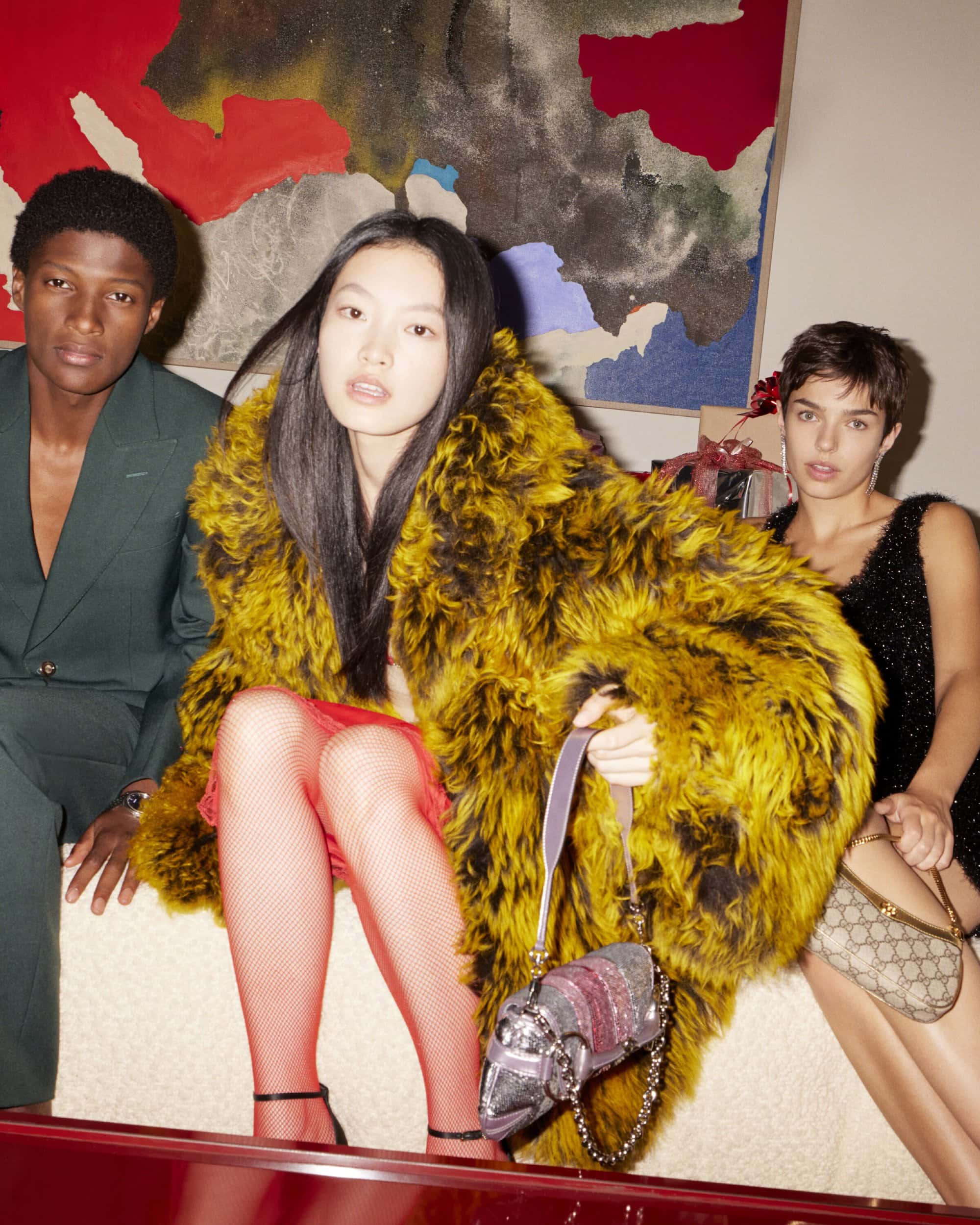 Three stylish fashion models pose indoors against a wall with an abstract painting, showcasing designer outfits and accessories.