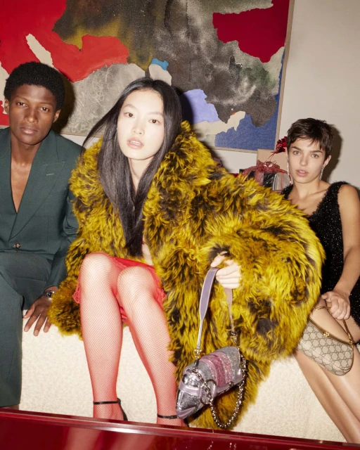 Three stylish fashion models pose indoors against a wall with an abstract painting, showcasing designer outfits and accessories.