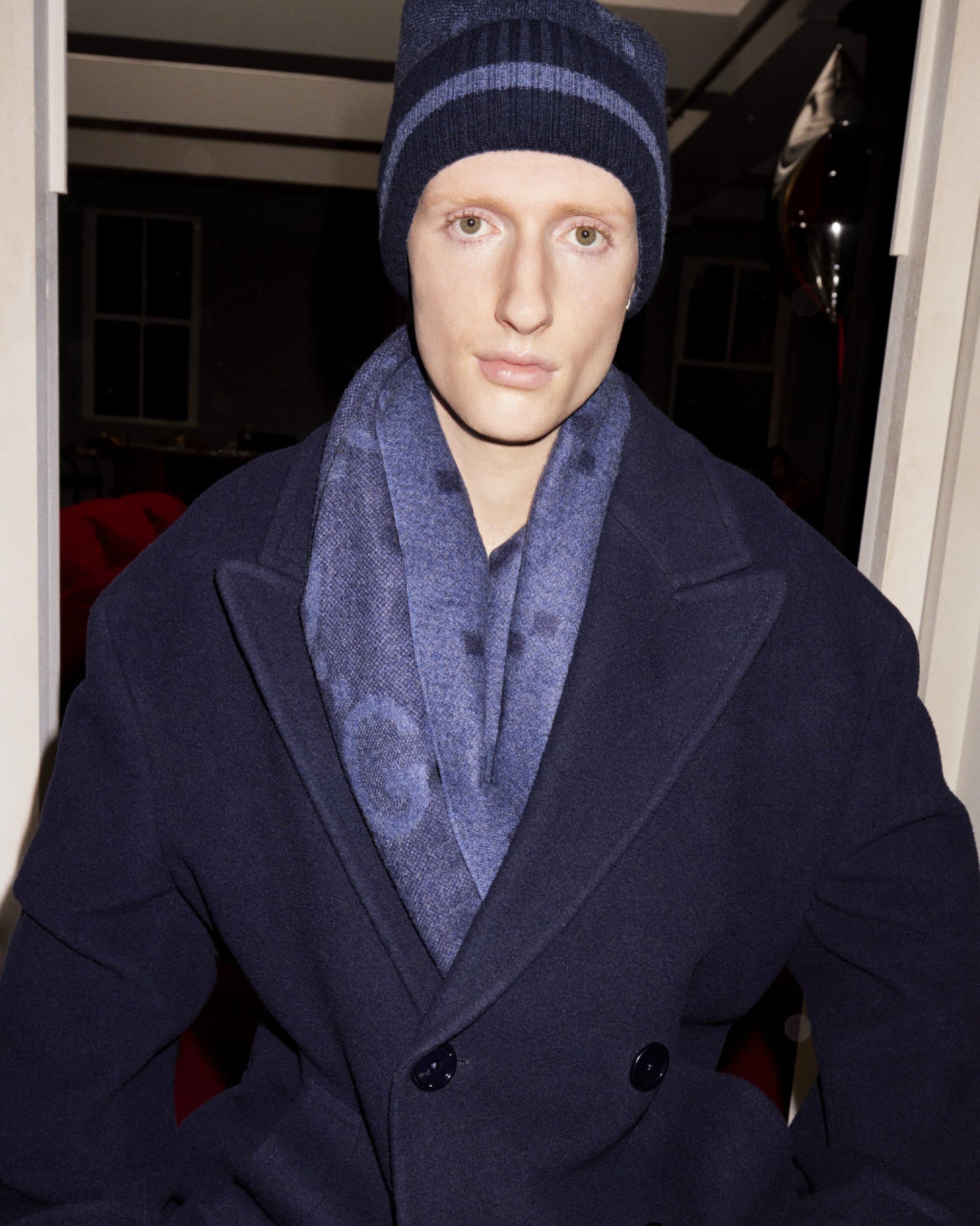 Adult male with fair skin wearing a navy blue beanie, scarf, and overcoat in an indoor setting with neutral facial expression.