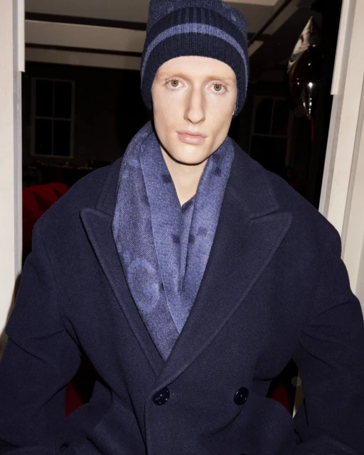 Adult male with fair skin wearing a navy blue beanie, scarf, and overcoat in an indoor setting with neutral facial expression.