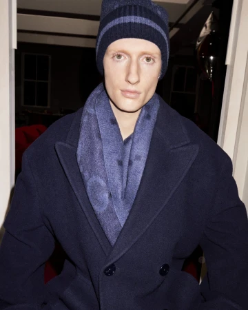 Adult male with fair skin wearing a navy blue beanie, scarf, and overcoat in an indoor setting with neutral facial expression.