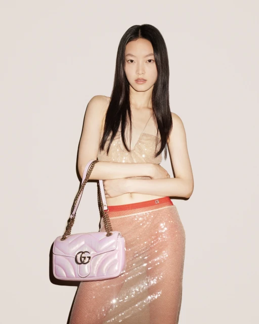 A stylish Asian model poses indoors with a pink Gucci purse, wearing a glittery top and sequined skirt against a light background.