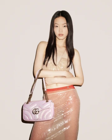 A stylish Asian model poses indoors with a pink Gucci purse, wearing a glittery top and sequined skirt against a light background.