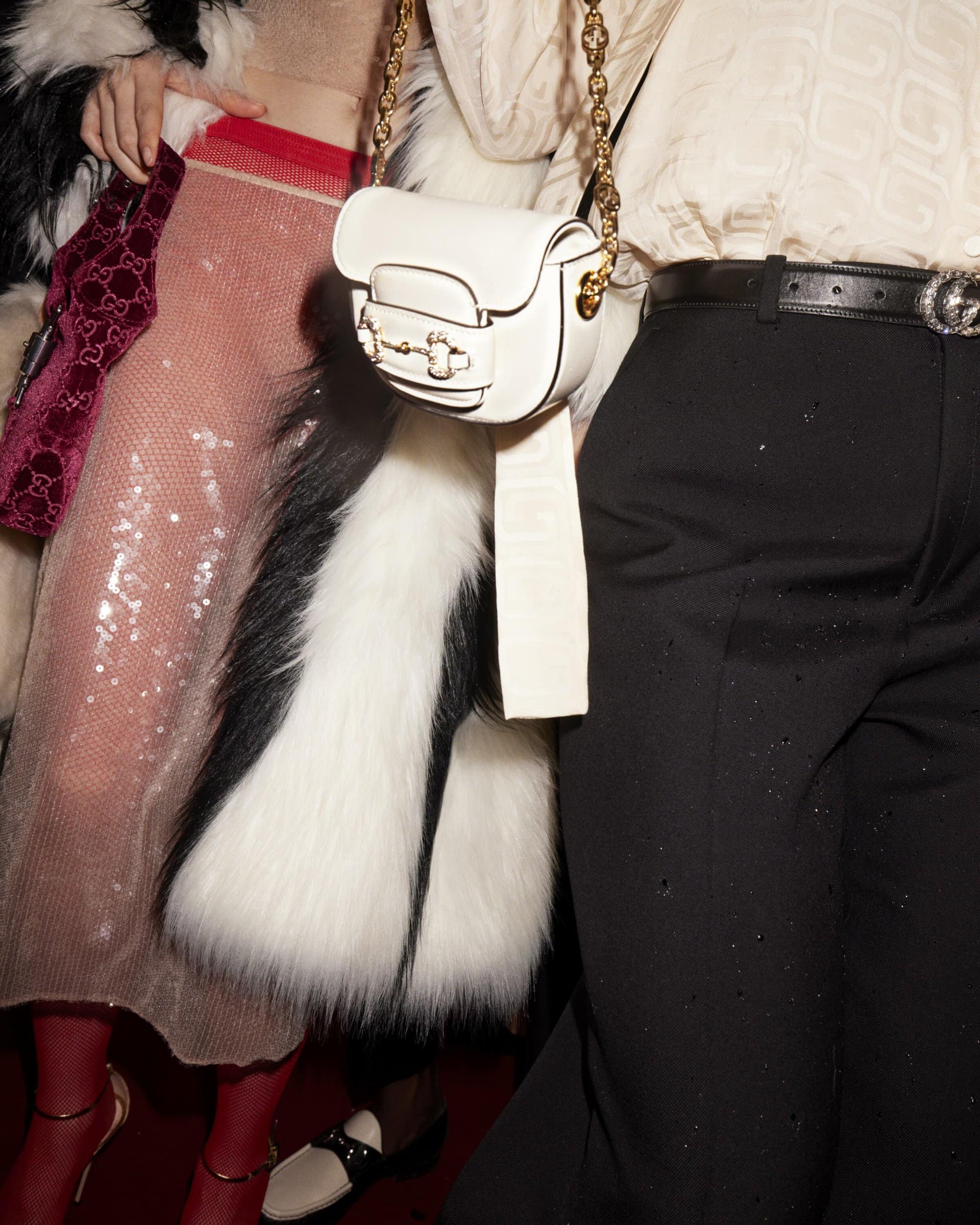 A close-up of two fashion models at an event, one with a white purse, black pants, and the other with a sparkly dress and fur coat.