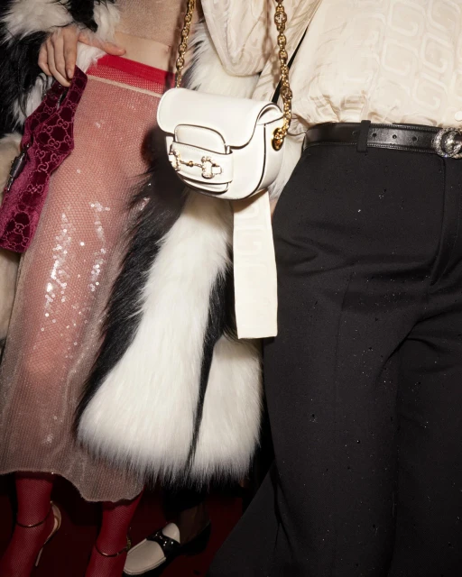 A close-up of two fashion models at an event, one with a white purse, black pants, and the other with a sparkly dress and fur coat.