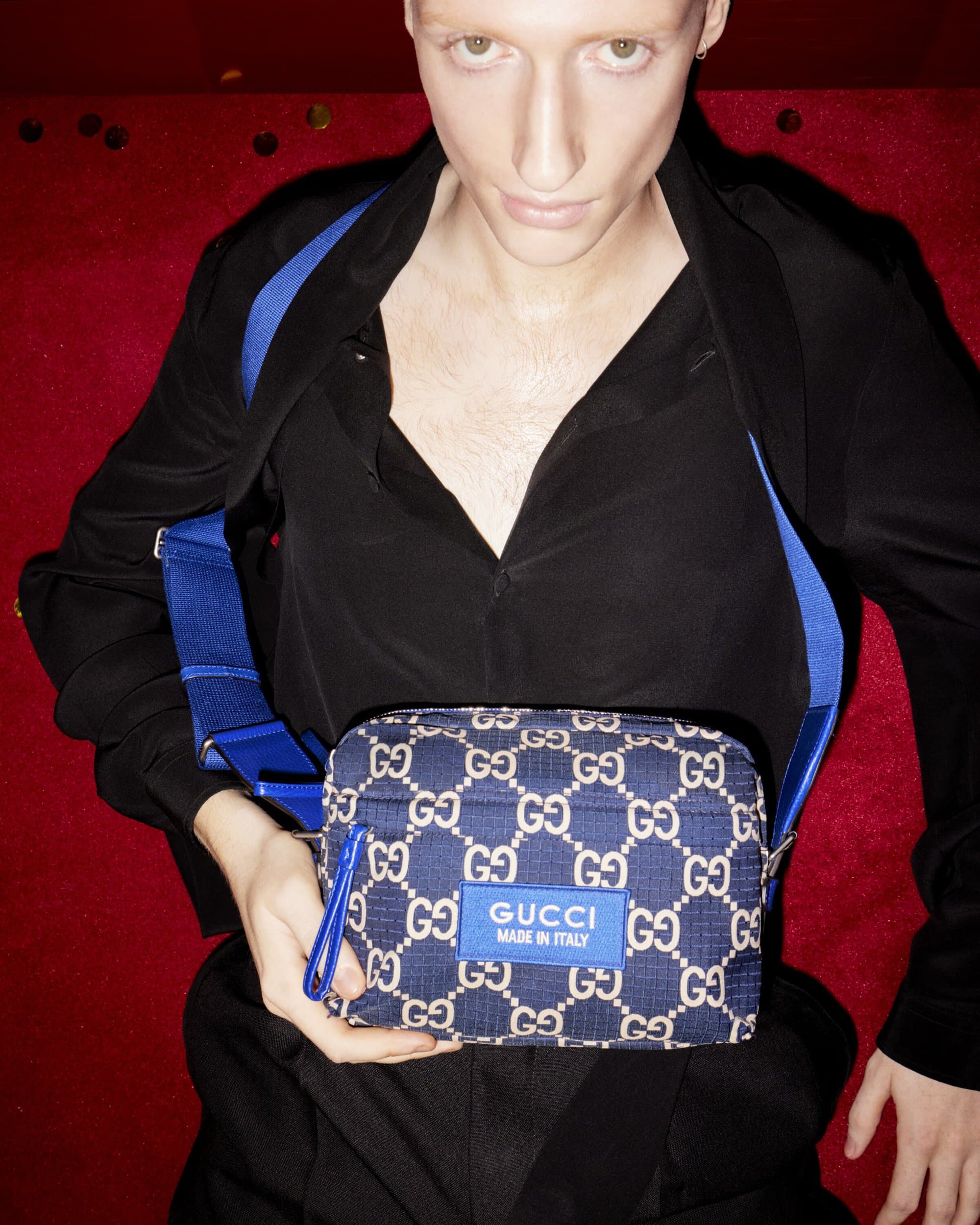 Person with blue patterned Gucci purse lying on red surface. Model with fashion accessory in a studio setting with warm, moody lighting.