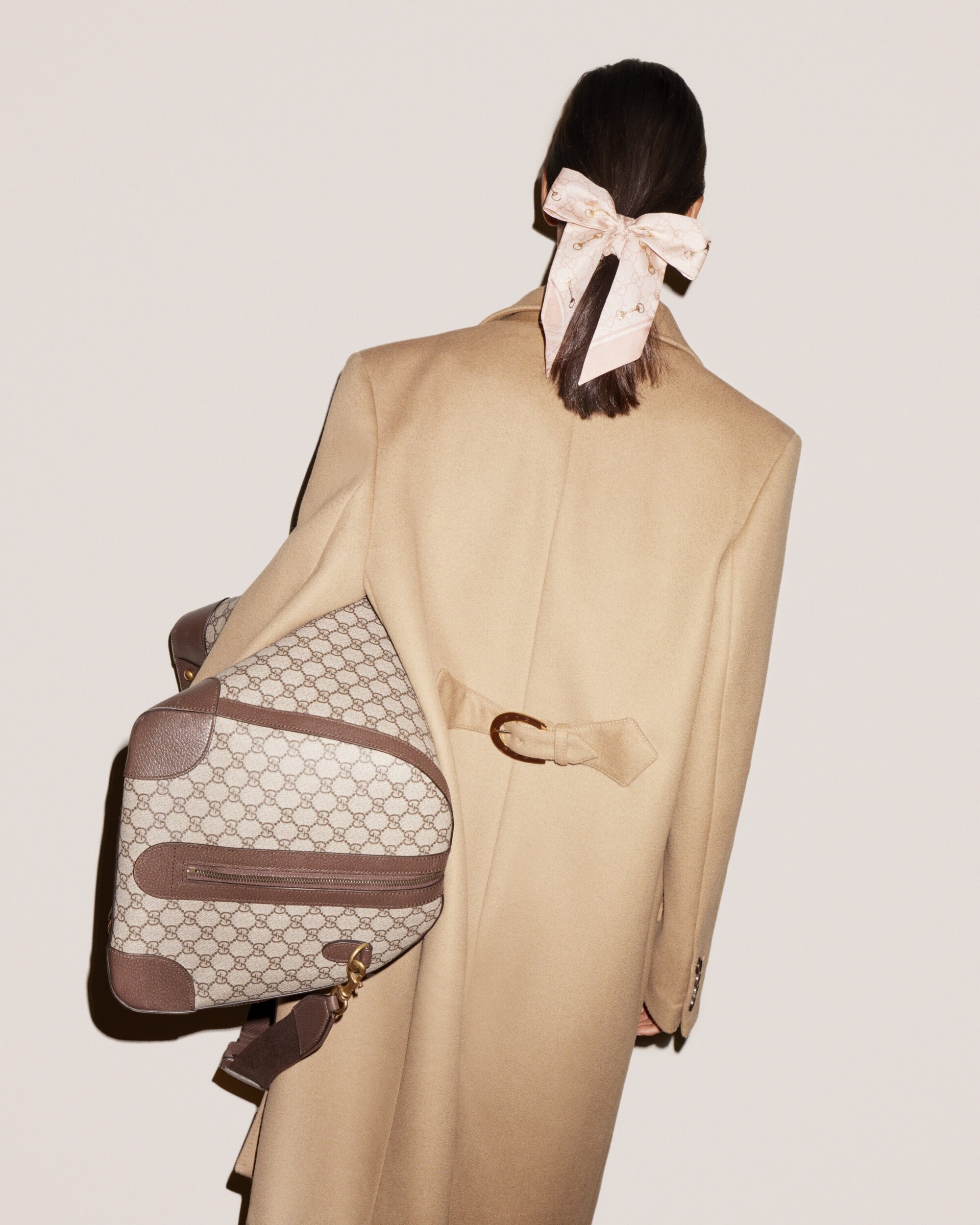 Woman with her back turned wearing a tan coat, holding a patterned duffel bag, and a hair ribbon posing in front of a plain wall.