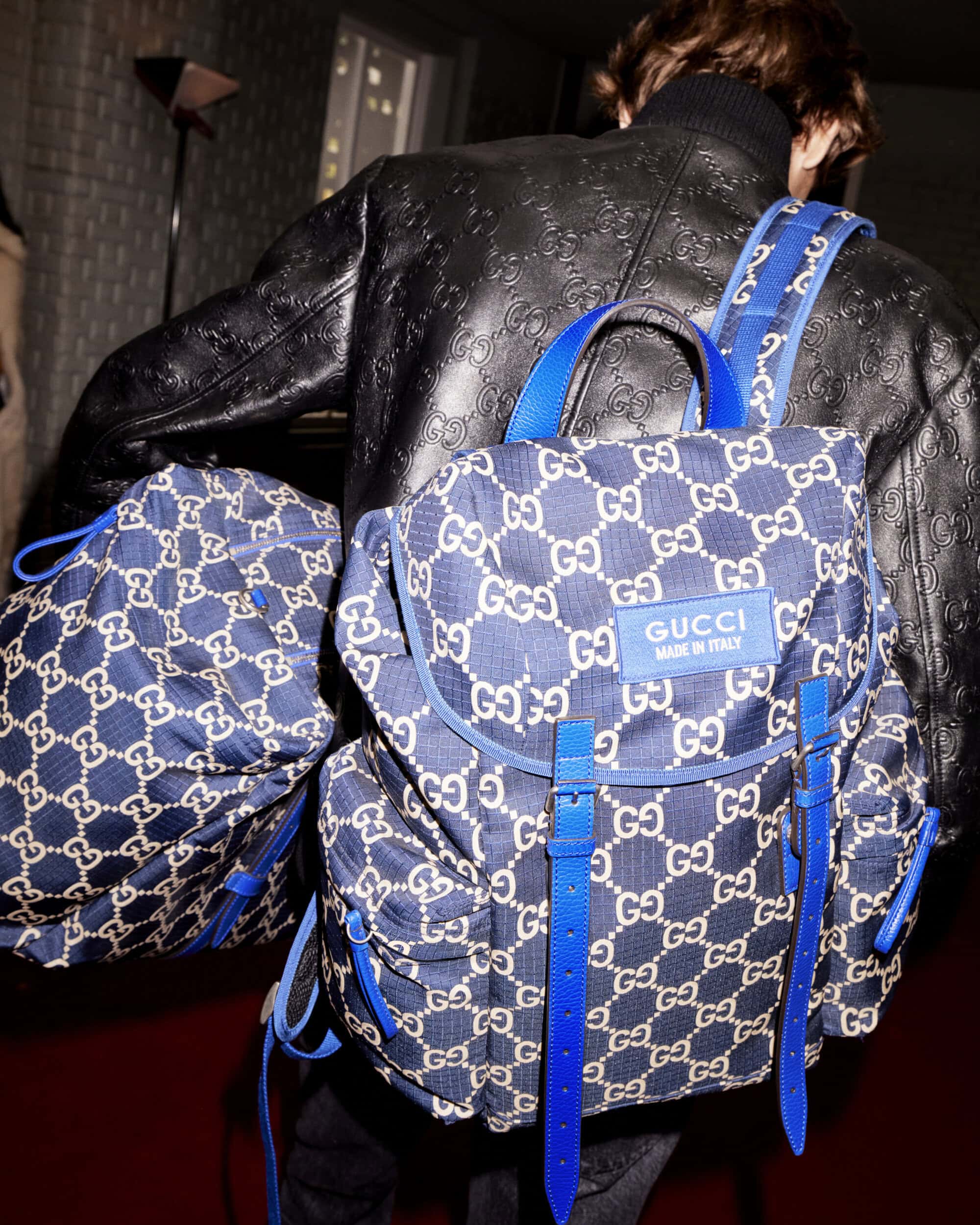 Person from behind wearing a black jacket and a Gucci backpack in blue and white with blue straps and accents, indoor setting.