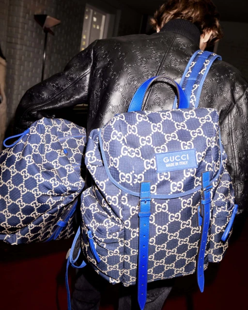 Person from behind wearing a black jacket and a Gucci backpack in blue and white with blue straps and accents, indoor setting.