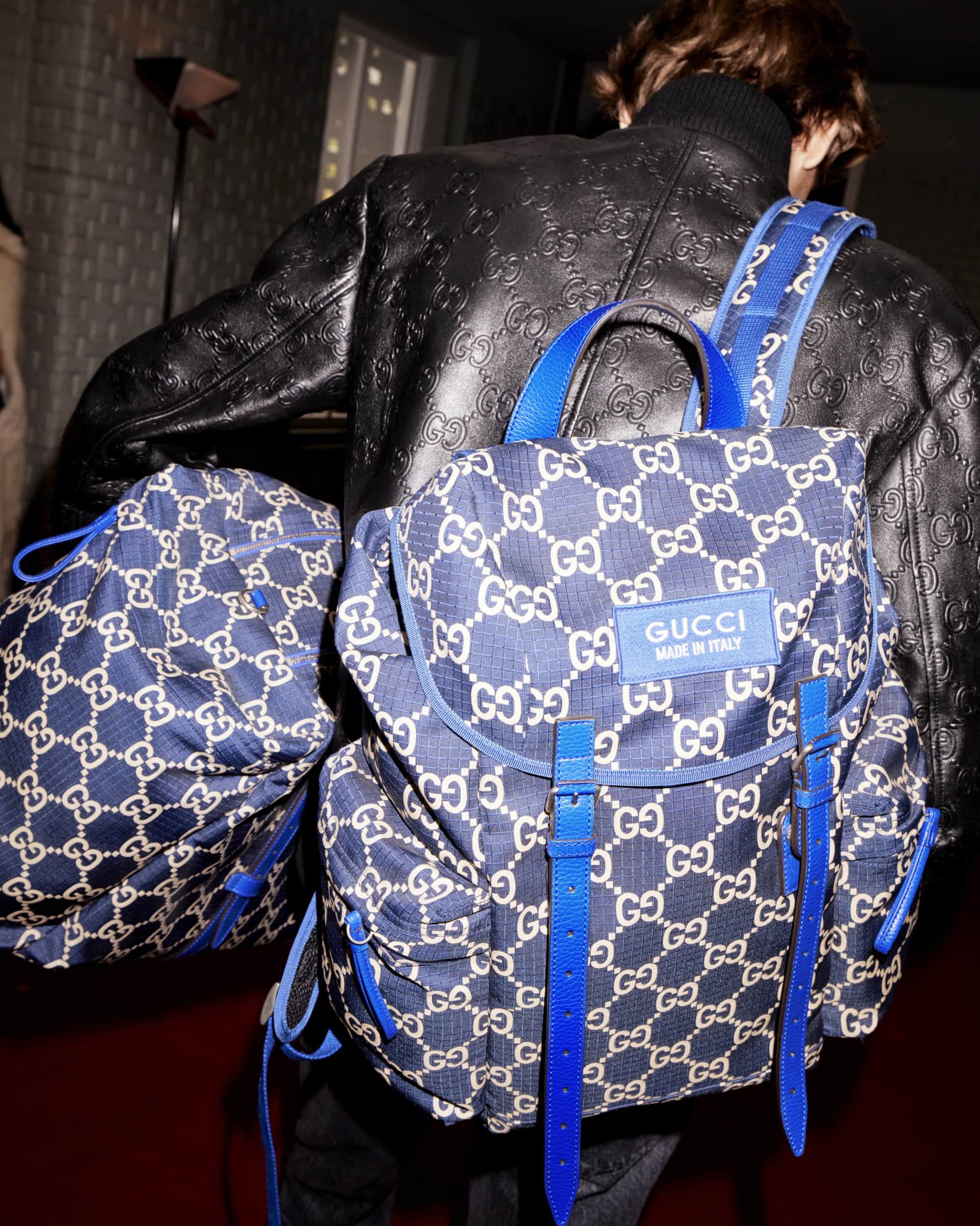 Person wearing a black leather jacket with two blue Gucci backpacks. One backpack is worn on their back, and another is on their side.