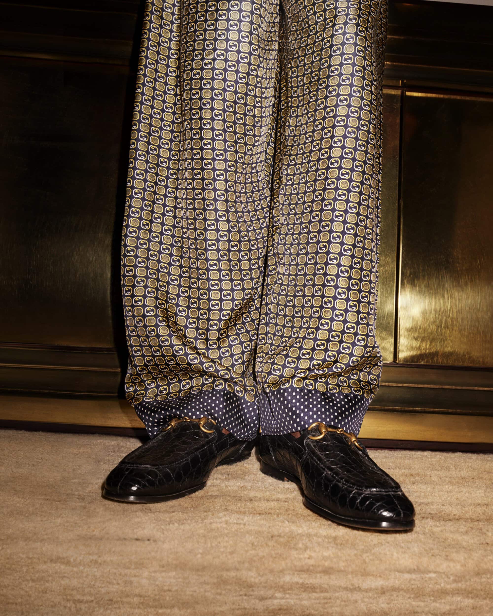 Close-up of patterned silk pants with a polka dot cuff and black crocodile loafers with metal buckle hardware on a carpet.