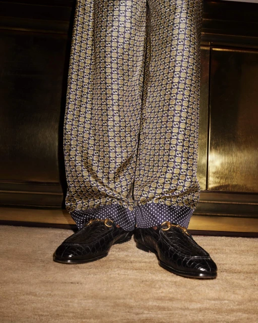 Close-up of patterned silk pants with a polka dot cuff and black crocodile loafers with metal buckle hardware on a carpet.