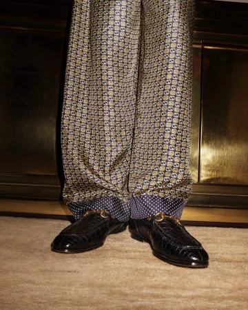 Close-up of patterned silk pants with a polka dot cuff and black crocodile loafers with metal buckle hardware on a carpet.