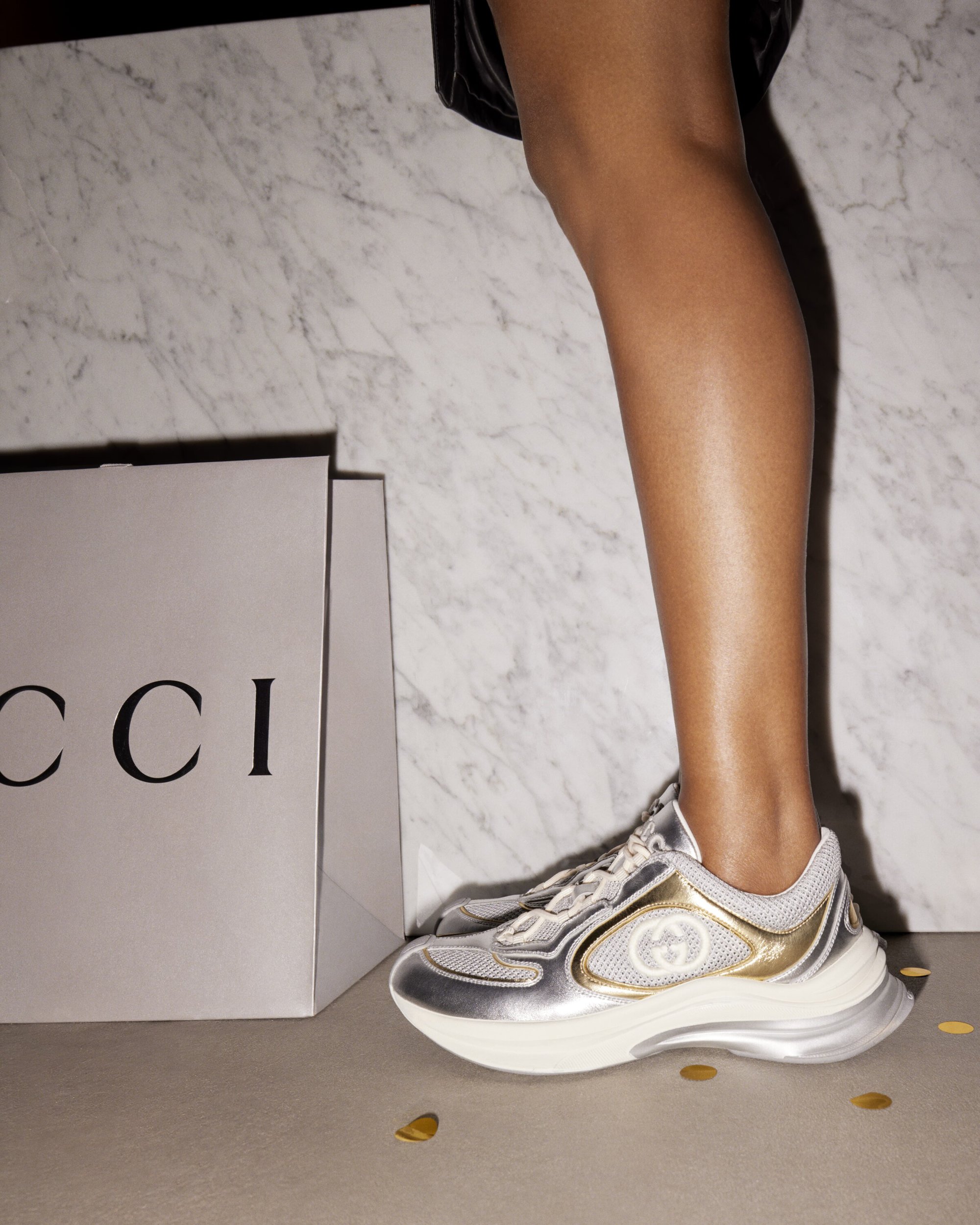Woman's leg and foot wearing a metallic Gucci sneaker near a Gucci shopping bag, with gold confetti on the floor and a marble background.
