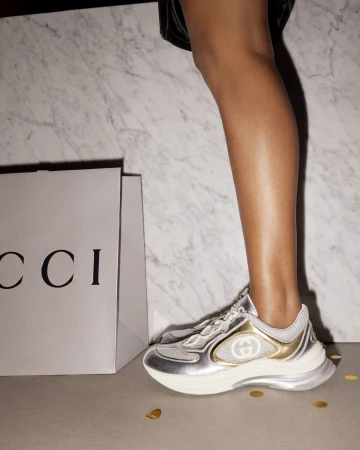 Woman's leg and foot wearing a metallic Gucci sneaker near a Gucci shopping bag, with gold confetti on the floor and a marble background.