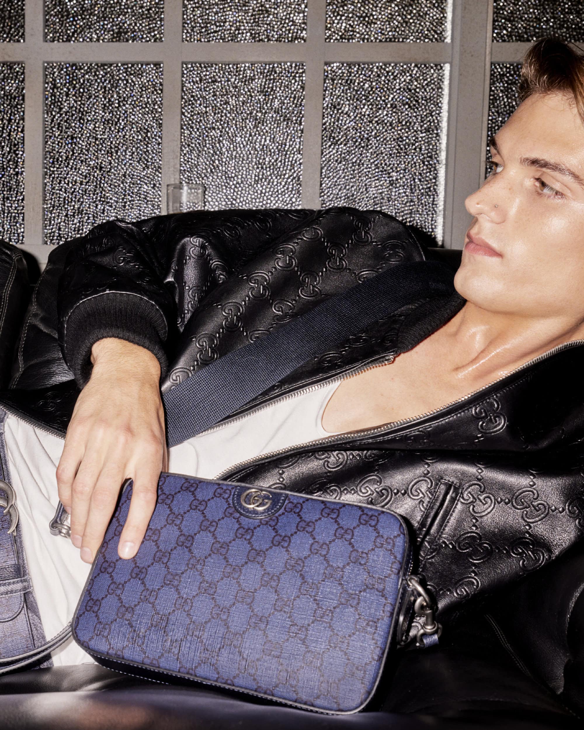 A fair-skinned adult male poses with a Gucci-patterned blue bag. He wears a black leather jacket and a white undershirt, indoors.