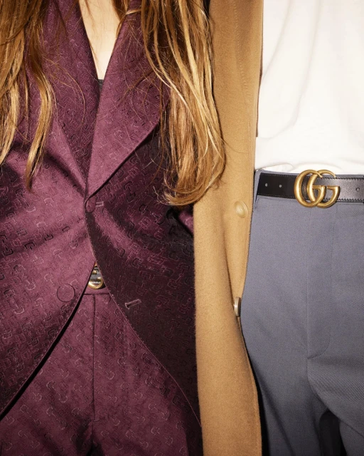 Two people wearing fashionable attire; one in a purple suit, the other in a tan coat, white shirt, and grey pants with a logo belt.