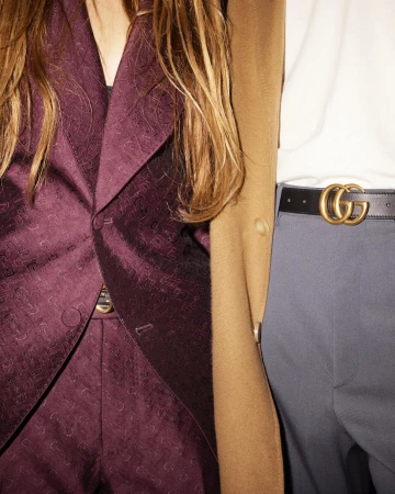 Two people wearing fashionable attire; one in a purple suit, the other in a tan coat, white shirt, and grey pants with a logo belt.