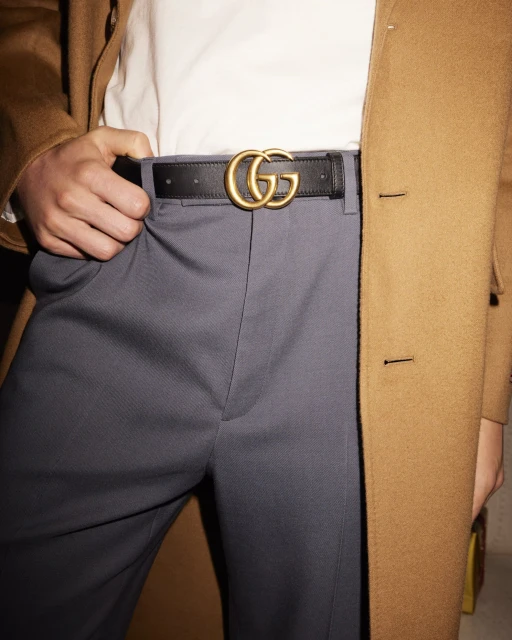 Close-up on a person wearing gray trousers, a white shirt, a brown coat, and a black leather belt with a gold-colored logo buckle.