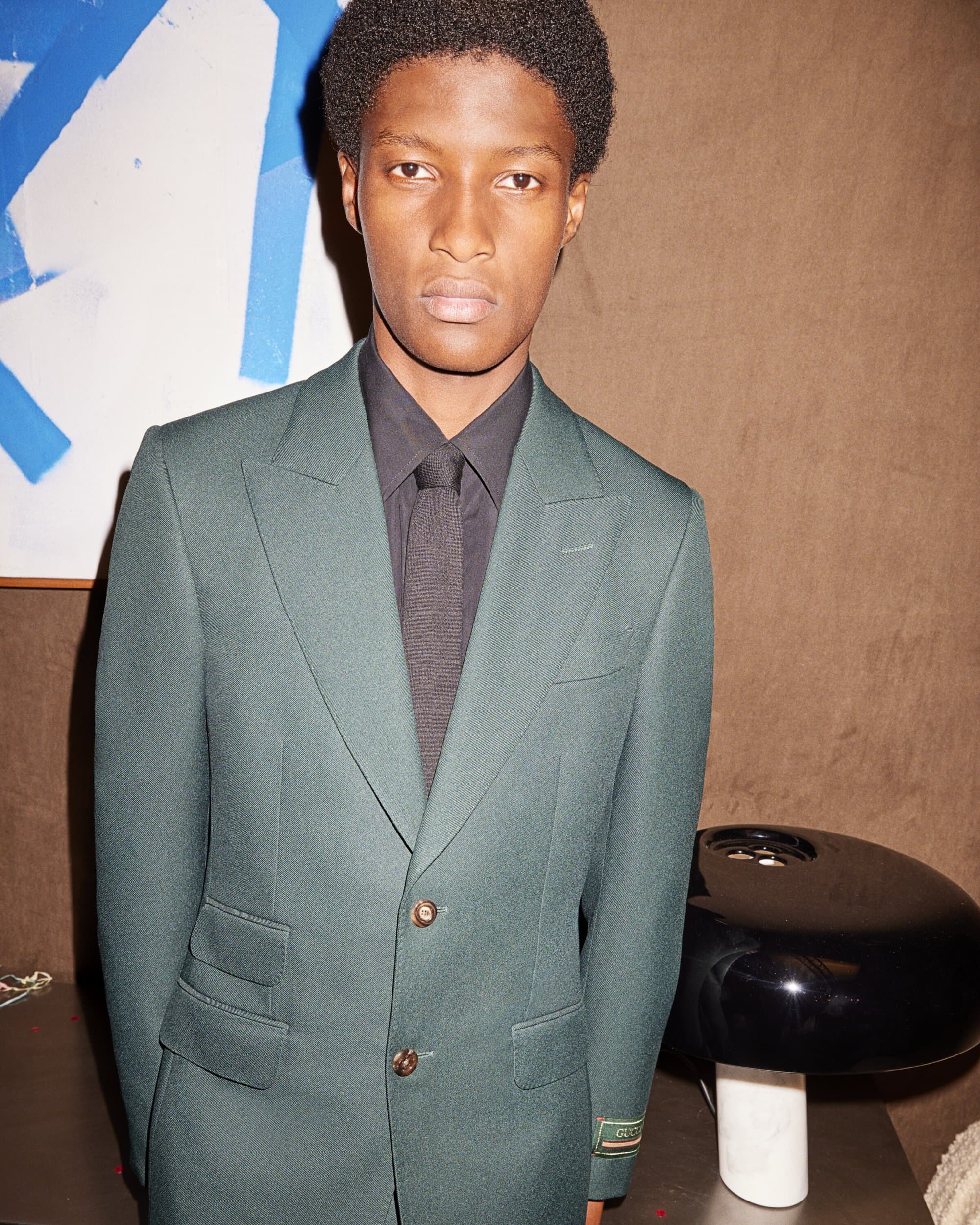 Portrait of a Black male model in a green suit and black shirt, posed in front of abstract art in an indoor setting, fashion editorial.