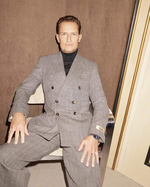 Mature man in grey herringbone suit and black turtleneck, seated indoors against brown background. He wears a ring and watch.