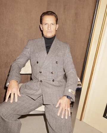Mature man in grey herringbone suit and black turtleneck, seated indoors against brown background. He wears a ring and watch.