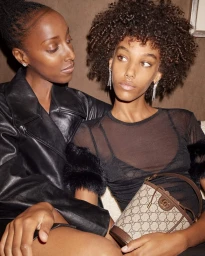 Two women seated together, one in a black leather jacket and one in a sheer black top holding a monogram bucket bag.