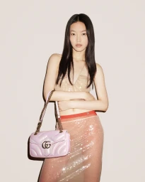 Model with long dark hair wearing a sequined top and pink skirt holding a quilted light pink shoulder bag against a neutral backdrop