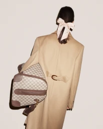 Model from behind wearing a camel coat and hair bow holding a monogram duffel bag against white seamless backdrop