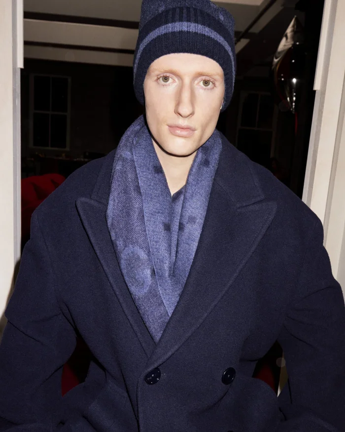 Model in navy blue coat, scarf, and matching beanie against an interior background for Gucci Holiday 2023 campaign.