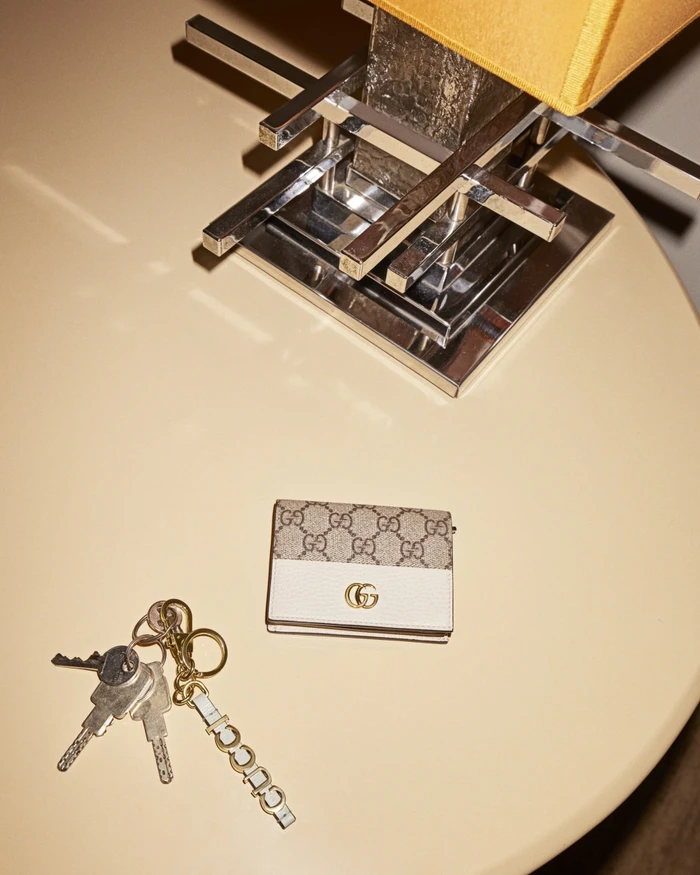 Gucci monogram wallet and keychain on a round beige table next to a geometric chrome lamp