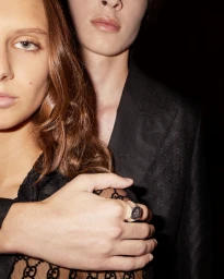 Close-up portrait of two models wearing black Gucci apparel with a hand wearing a signet ring visible in the foreground