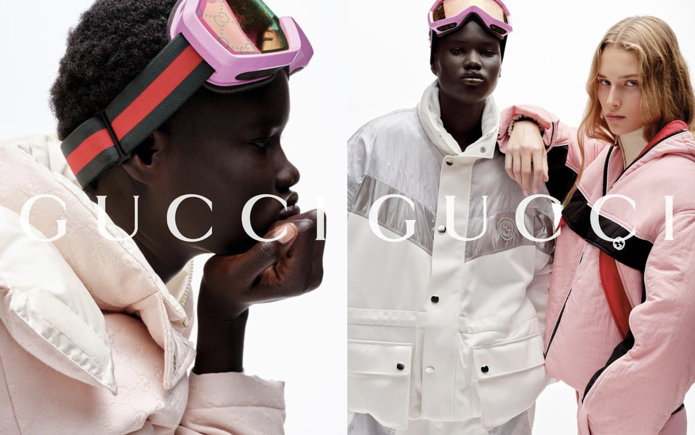 Two fashion models pose in a Gucci winter apparel ad. One wears pink ski goggles and a jacket, the other in white, on a white backdrop.