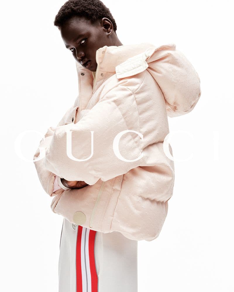 Fashion shot of a dark-skinned model wearing a light pink puffer jacket with a hood and white pants with red stripes, on a white background.