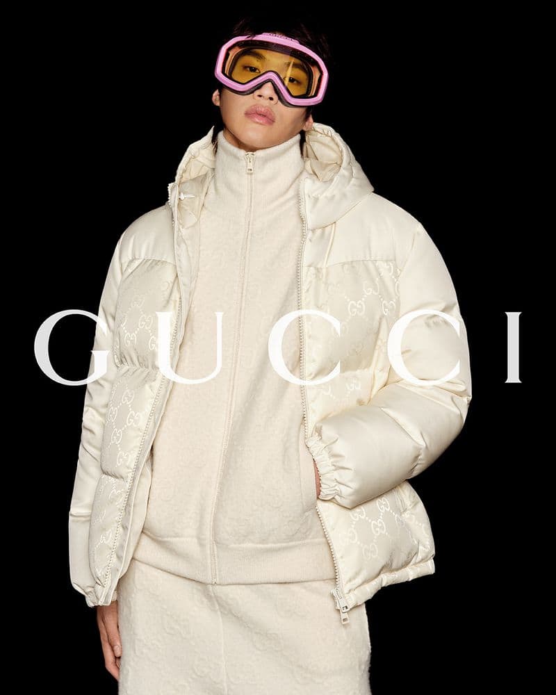 Person wearing white puffer jacket, turtleneck, and pink ski goggles against a black backdrop, with Gucci logo overlayed.