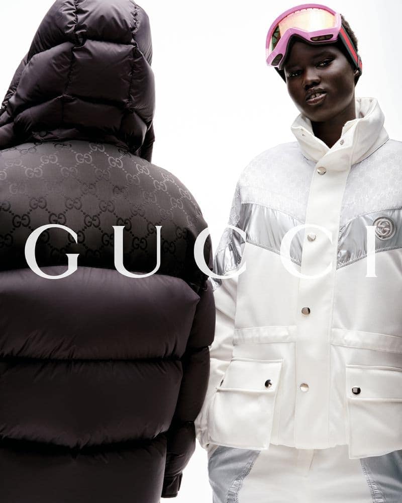 Two people wearing black and white Gucci winter coats. The woman on the right wears pink ski goggles and stands on a white backdrop.