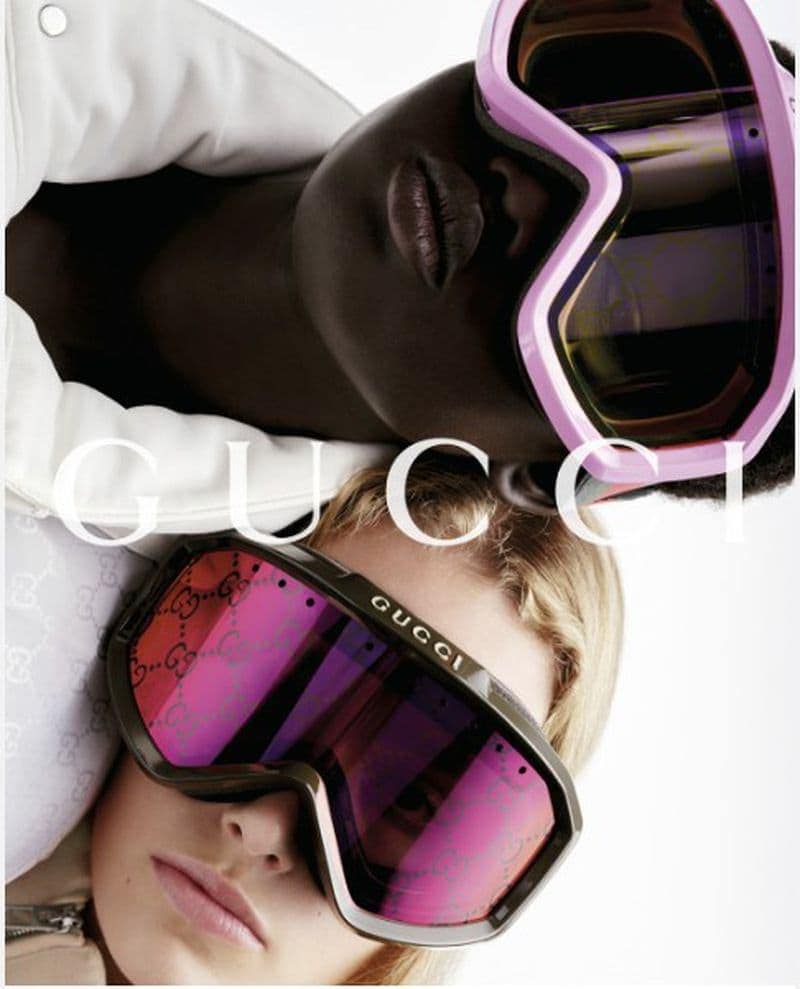 Fashion shot of two women wearing Gucci ski goggles. One is blonde and the other is Black. The background is light, and the image is a close-up.