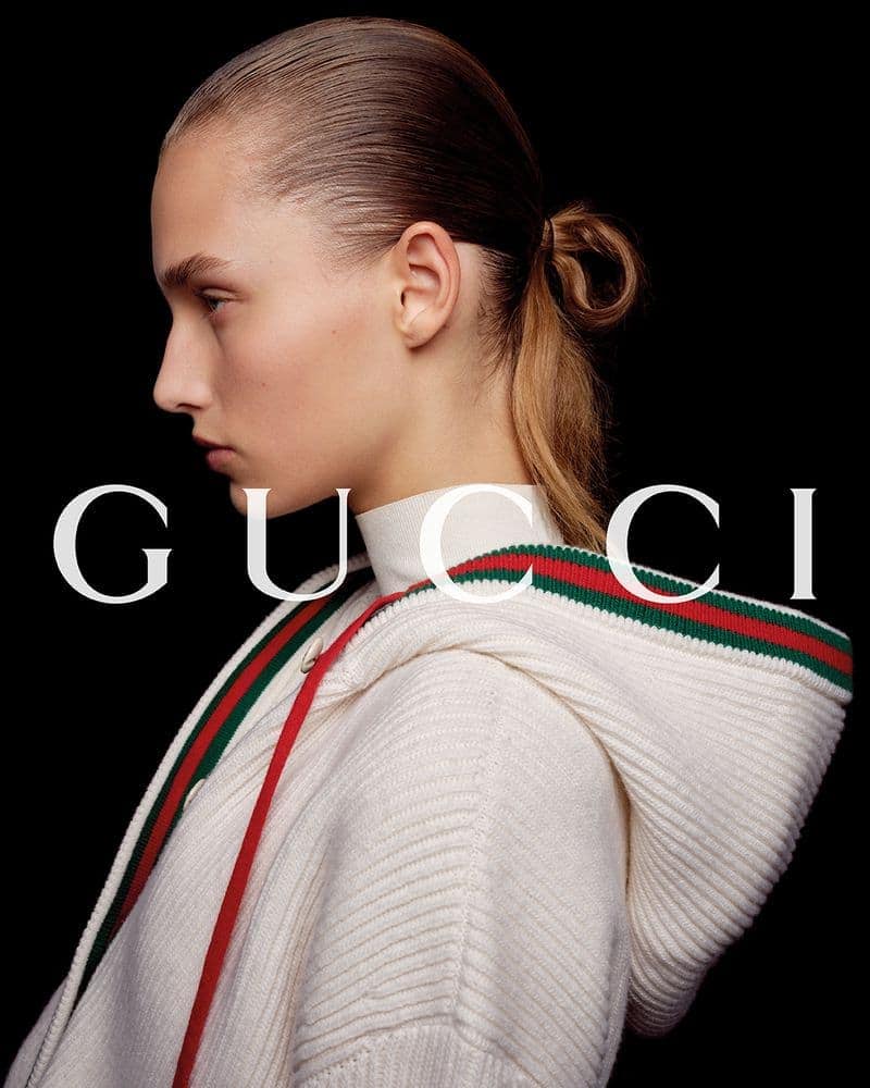 Profile portrait of a young fair-skinned woman in white hooded sweater, against black background, featuring a prominent Gucci logo.