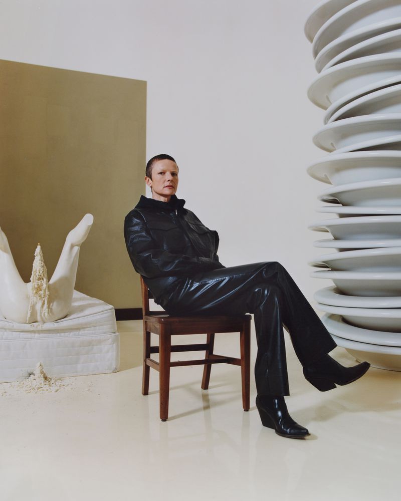 Person in black leather outfit sitting on a wooden chair in a minimalist studio setting with art installations and neutral backdrop.