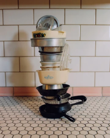 Stack of cookware includes mixing bowls, glass lids, frying pans and baking pans, stacked unevenly on a kitchen counter with tile backsplash.