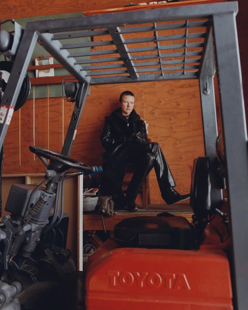 Adult with short, red hair and black clothing seated inside the frame of a forklift, in front of a wooden paneled wall.