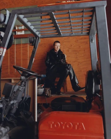 Adult with short, red hair and black clothing seated inside the frame of a forklift, in front of a wooden paneled wall.