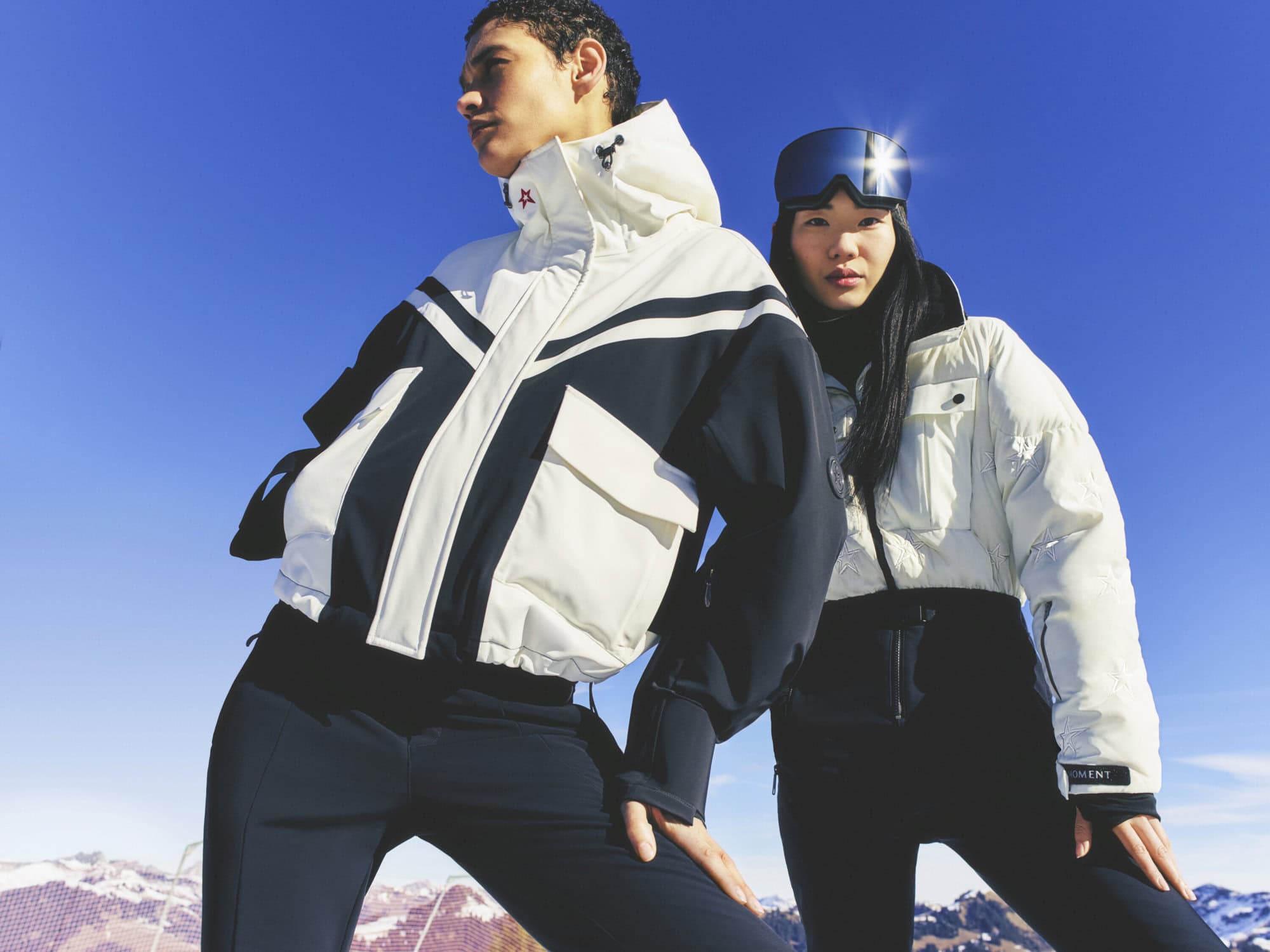 Two models showcase winter fashion in a snowy mountain setting under a bright blue sky, wearing puffer jackets and ski goggles. 