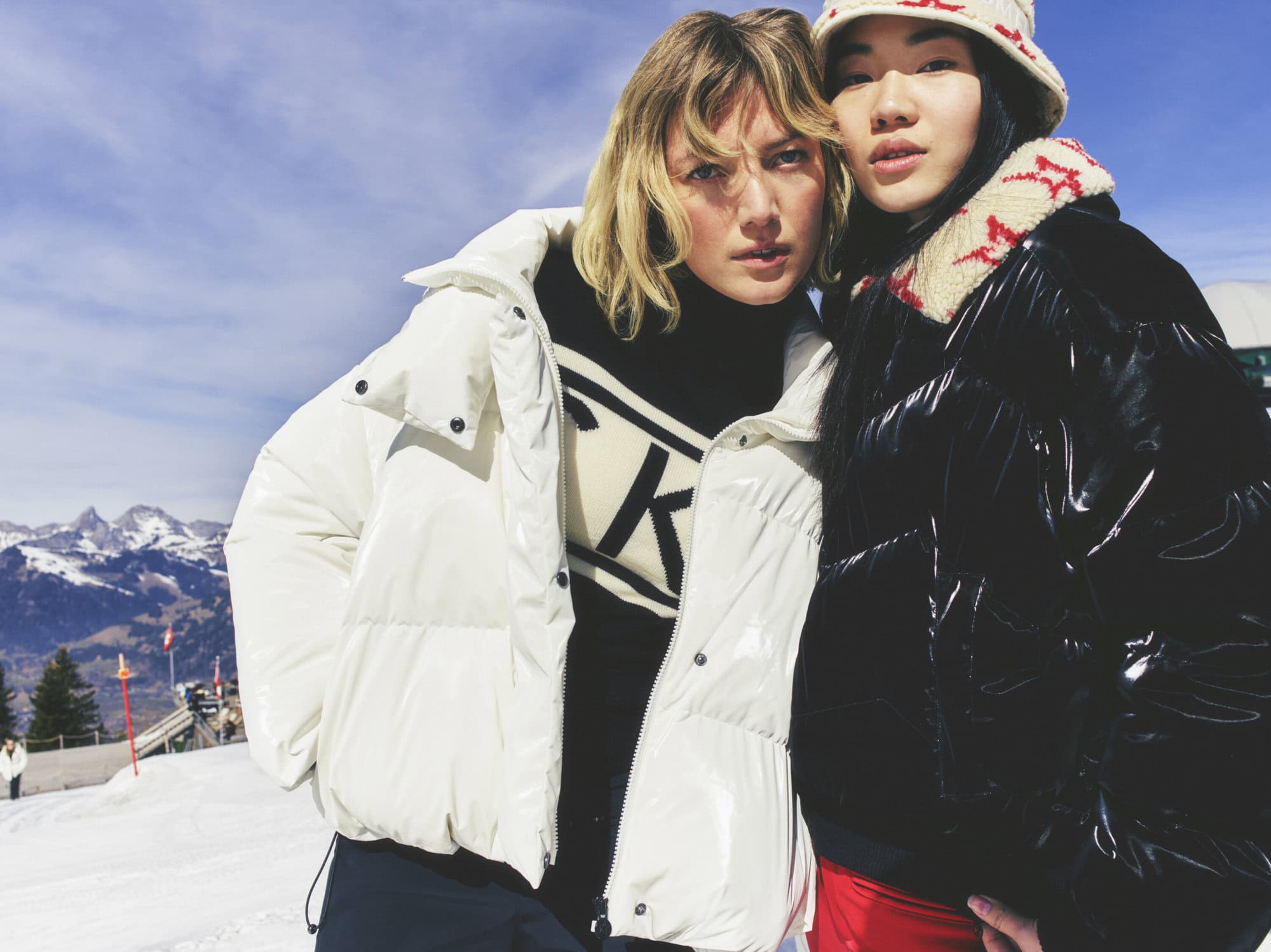 Two women pose in winter clothing against a snowy mountain backdrop. One wears a white puffer jacket; the other, a black jacket and hat.