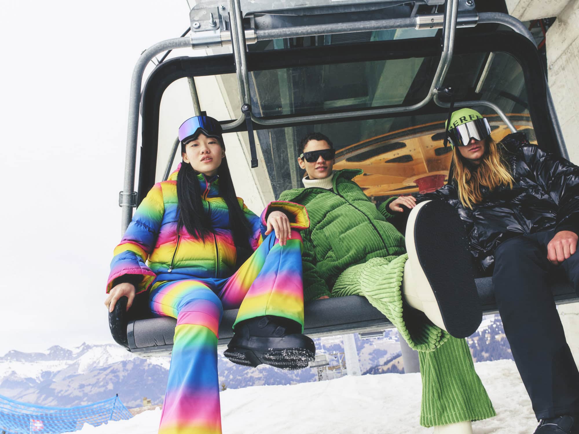 Three people wearing colorful ski suits sit in a chair lift at a ski resort surrounded by snow-covered mountains on a bright day.